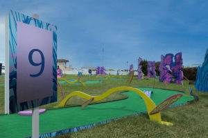 Sign indicating Hole 9 in front of the Adventure Golf and Sports MiniLinks™ hole with various obstacles including a bridge and a yellow curved bar above the green.