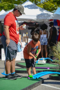 On an AGS SplitShot® blue bridge, a young boy attempts to knock his mini golf ball across it to the next green while his father watches from behind.