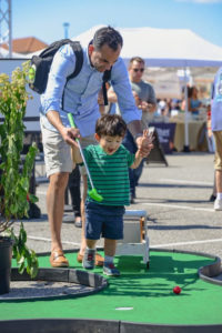 Father helping smiling young boy chase after his mini golf ball on the portable putting green at the Famous Food Festival held at Tanger Outlets