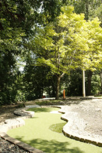 Pulled back image of one of the putting green, bricks framing it down each side and surrounded by bright green trees, at Veterans Memorial Park.