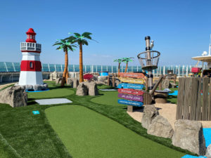 With the ocean in the background, a mini golf hole is installed on RCL Liberty Cruise Ship with theme elements such as a miniature lighthouse, palm trees, and pirate ship’s crows nest.