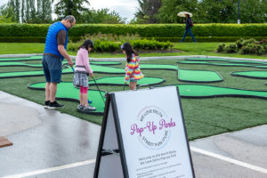 A man and two young girls putting in the rain on an AGS mini golf course at a municipal pop-up park