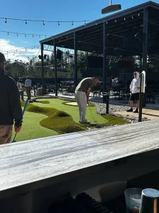 Player teeing off on the first mini golf hole faces faux sand trap and rough turf on way to the hole cup