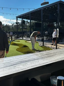 Player teeing off on the first mini golf hole faces faux sand trap and rough turf on way to the hole cup